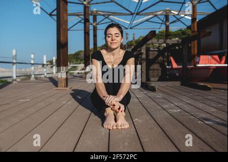 Lächelnde Frau, die auf Holzboden am Meer sitzt und die Sonne genießt, nachdem sie Sport gemacht hat. Stockfoto