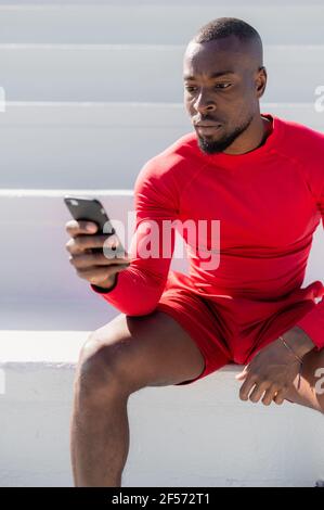 Vertikale Nahaufnahme von schwarzen afrikanischen muskulösen Mann mit Sportswear sitzen auf weißen Stufen mit ernsthaftem Gesicht mit Smartphone. Stockfoto