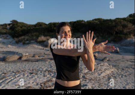 Junge Frau in Sportbekleidung tun Stretching-Übungen für Hals und Arme an der Küste mit entspanntem Smiley nach vorne schauen Gesicht beim Atmen frisch A Stockfoto
