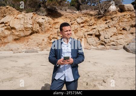 Latein Mann im eleganten Anzug am Strand mit Smartphone und wegschauen. Entfliehen Sie der Stadt auf der Suche nach einem friedlichen Ort Stockfoto