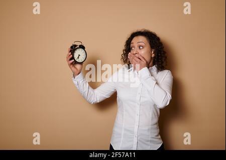Eine betäubt hispanische lockige Frau hält einen Wecker in ihren Händen und schaut ihn mit Entsetzen an, bedeckt ihren Mund mit ihrer Hand. Die Zeit ist flüchtig. Concep Stockfoto
