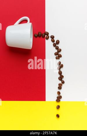 Kaffeebohnen, die aus einer weißen Tasse auf weißem, rotem und gelbem Hintergrund verschüttet werden. Stockfoto