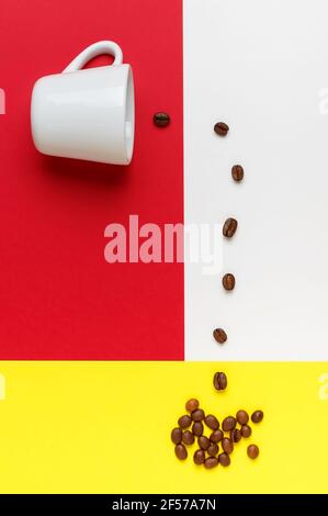 Kaffeebohnen, die aus einer weißen Tasse auf weißem, rotem und gelbem Hintergrund verschüttet werden. Stockfoto