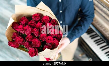 Der Bräutigam im Anzug mit Hochzeitsstrauß steht neben dem Klavier. Romantischer Kerl gibt einen luxuriösen Strauß roter Rosen zu seiner Freundin auf einem Datum für Stockfoto