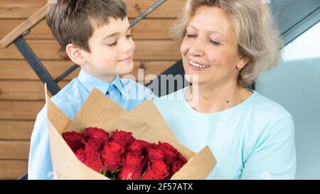Ein süßer Junge in einem blauen Hemd verleiht seiner Großmutter einen luxuriösen Strauß roter Rosen zu ihrem Geburtstag, Muttertag, Jubiläum. Herzliche Familie relat Stockfoto