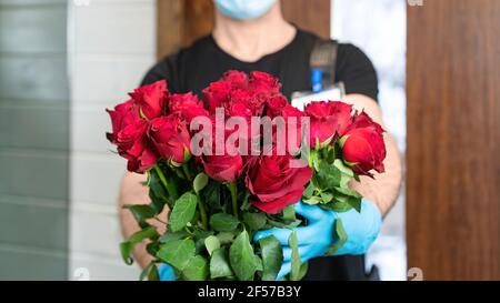 Blumenlieferservice. Ein Kurier mit medizinischer Maske und Handschuhen ermöglicht die kontaktlose Lieferung eines luxuriösen Straußes roter Rosen während des COVID-1 Stockfoto