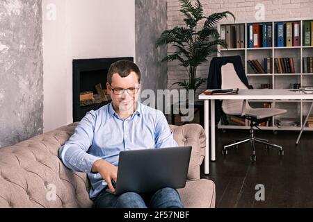 Ein junger erfolgreicher Mann in einem Hemd und einer Brille lächelt und schaut in einen Laptop, der auf einer Couch in seinem Heimbüro liegt. Das Konzept des Erfolgs, Remote-Arbeit und Stockfoto