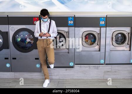 Selektiver Fokus eines jungen Mannes, der eine Maske trägt und Hosenträger, die Musik von einem Mobiltelefon und Kopfhörer hören Beim Warten auf die Waschmachi Stockfoto