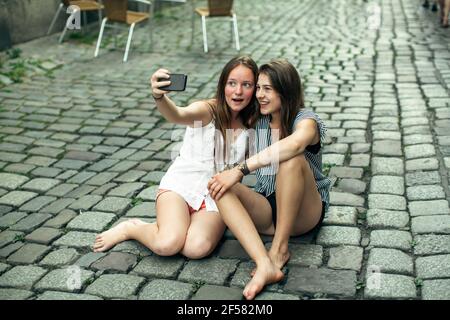 Zwei Freundinnen machen ein Selfie auf einem Smartphone, das auf dem Bürgersteig in der Altstadt sitzt. Stockfoto