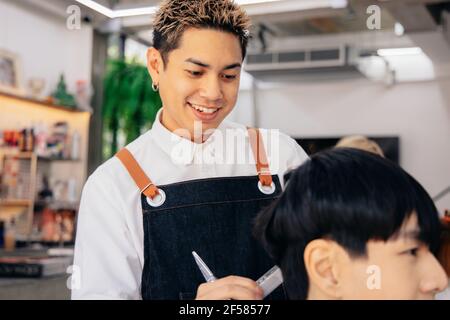 Glücklich junge asiatische männliche Friseur lächelnd trägt Schürze in modernen Salon hält Pinsel und Schere Schneiden Haar des Kunden Stockfoto