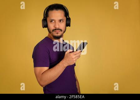 Junger Mann, der mit Kopfhörern Musik hört und mit dem Smartphone spielt. Gelber Hintergrund. Stockfoto