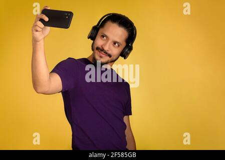 Junger Mann nimmt ein Selfie und hört Musik mit Kopfhörern und spielt mit dem Smartphone. Gelber Hintergrund. Stockfoto