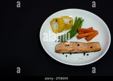 Lachssteak gebraten mit Gemüse, gegrilltem Mais, Spargel, Karotte, Pfeffer auf schwarzem Hintergrund. Stockfoto
