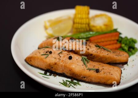 Zwei Stück Lachssteak mit Gemüse, gegrilltem Mais, Spargel, Karotten, grünen Erbsen, Zitrone, Pfeffer auf schwarzem Hintergrund. Stockfoto