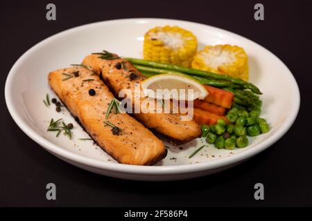 Zwei Stück Lachssteak mit Gemüse, gegrilltem Mais, Spargel, Karotten, grünen Erbsen, Zitrone, Pfeffer auf schwarzem Hintergrund. Stockfoto