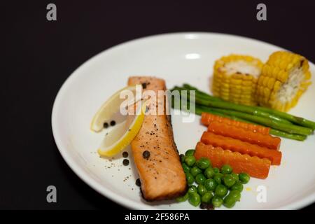 Lachssteak mit Gemüse, gegrilltem Mais, Spargel, Karotten, grünen Erbsen, Zitrone, Pfeffer auf schwarzem Hintergrund. Stockfoto