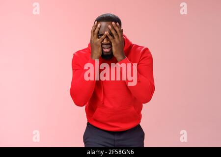 Schockiert aufgeregt jungen afroamerikanischen bärtigen Kerl in roten Kapuzen Facepalm verstecken Gesicht Hände gucken durch die Finger traurig verwüstet verlieren Hoffnung, traurig stehen Stockfoto