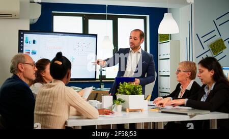 Manager zeigt auf digitales, interaktives Whiteboard mit Wachstumsanalyse, Diagrammen, Statistiken und Daten. Verschiedene Personen, die während einer Konferenz im Broadroom arbeiten und über Unternehmensprobleme sprechen. Stockfoto