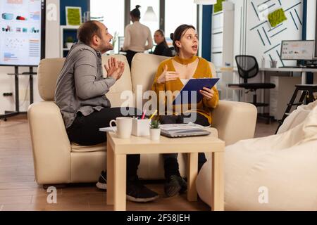 Verärgerter Mann Manager argumentiert Frau Angestellter für schlechte Arbeitsergebnisse, sitzen auf der Couch, während verschiedene Kollegen arbeiten Angst im Hintergrund. Geschäftsfrau Angestellter wütete über Multitasking schwierige Arbeit. Stockfoto