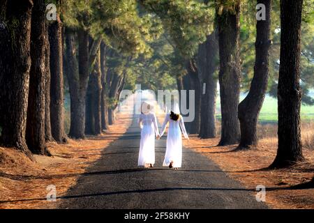 Pleiku Stadt, Gia Lai Provinz, Vietnam - 7. März 2021: Zwei Mädchen in traditioneller Kleidung ist auf der Straße mit zwei Reihen von Pinien, die p zu Fuß Stockfoto