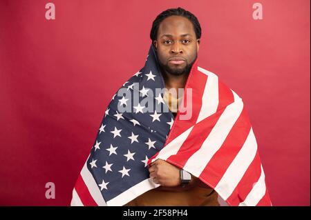 Selbstbewusster afroamerikanischer Mann Kämpfer für Rechte stehen mit US-Flagge bedeckt, leiden an Ungerechtigkeit, protestieren gegen Rassismus, isoliert auf roter Wand, schwarze Leben Materie Konzept Stockfoto