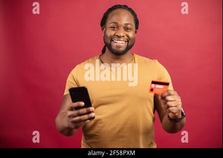 Afroamerikanischer Mann, der isoliert auf rotem Wandhintergrund steht, aufgeregt für zukünftige Einkäufe, Geld ausgeben, Kreditkarte und Handy halten, Debitkarte für die Zahlung von Versorgungsunternehmen oder Rechnungen verwenden Stockfoto