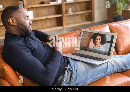 Ein afroamerikanischer Mann trifft sich virtuell mit einer Freundin, Freundin oder Kollegin. Ein multirassischer Kerl verwendet Laptop für Videoanruf, legt auf Couch und spricht entspannt Stockfoto