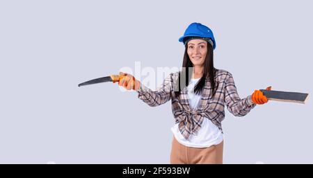 Geschlechterstereotypen brechen. Selbstbewusste Frau in einem blauen Schutzhelm mit einem Brett und einer Handsäge. Studio grauer Hintergrund. Stockfoto