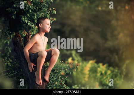 Ein Junge ohne Hemd sitzt auf einem teilweise gefällten Baum im Dorf. Stockfoto