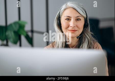 Porträt der schönen erfolgreichen selbstbewussten reifen grauhaarigen asiatischen Frau, Geschäftsfrau, Anwalt, Immobilienmakler, sitzt mit Kopfhörern vor dem Computer, Blick auf die Kamera, freundlich lächelnd Stockfoto