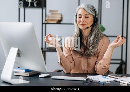 Entspannt ruhig schöne reife grauhaarige asiatische Frau, Geschäftsführerin, Call-Center-Betreiber, im Headset, in stilvoller Kleidung, Am Arbeitsplatz ausruhen, meditieren, Stress lindern, indem sie ihre Augen schließt Stockfoto