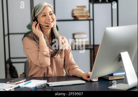 Selbstbewusste einflussreiche asiatische Senior-Frau, Geschäftsfrau, Manager, Callcenter-Betreiber, arbeitet im Büro, Kommuniziert per Videoanruf mit dem Kunden, führt Online-Beratung durch, nutzt Headset, lächelt Stockfoto