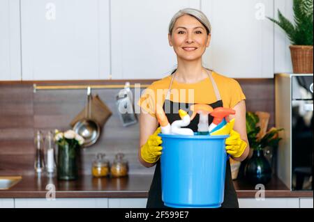 Freundliche reife grauhaarige asiatische Putzfrau oder Hausfrau in Handschuhen und eine Schürze steht in der Küche, hält große Eimer mit Waschmitteln und Lumpen, Blick auf die Kamera, lächelnd Stockfoto