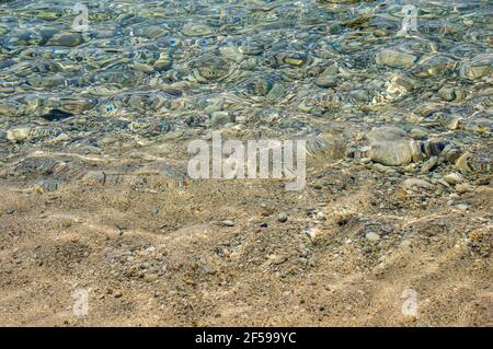 Nahaufnahme des kristallklaren Meerwassers mit Steinen in der Nähe des Strandes auf der griechischen Insel Korfu. Hintergrund mit Lichtbrechung auf transparenter Wasseroberfläche Stockfoto