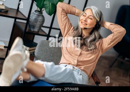 Entspannte schöne reife grauhaarige asiatische Geschäftsfrau, Freiberuflerin, sitzen im Büro, werfen ihre Beine auf dem Tisch, Pause während der Arbeit, Kreuzung ihre Arme hinter dem Kopf, lächelnd Stockfoto
