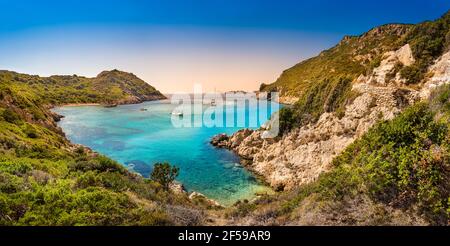 Boote in Bucht in der Nähe von Porto Timoni Strand auf Korfu Insel in Griechenland. Panorama der griechischen Küste mit grünen Bergen, klarem Meerwasser und Piratenbucht bei Sonnenuntergang Stockfoto