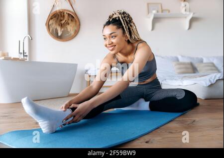 Fröhliche afroamerikanische junge Frau in Sportkleidung und mit Dreadlocks, kümmert sich um ihre Gesundheit, macht Fitness, führt einen gesunden Lebensstil, macht Stretching zu Hause auf der Matte, lächelt Stockfoto