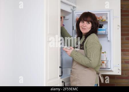 Junge Frau lächelt und öffnet den Kühlschrank, um die Zutaten zu bekommen. Garzeit. Stockfoto