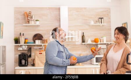 Mann jonglieren mit Orangen für die Frau, während die Zubereitung von leckeren und nahrhaften Smoothie. Gesunde unbeschwerte und fröhliche Lebensweise, Ernährung und Vorbereitung Frühstück in gemütlichen sonnigen Morgen Stockfoto