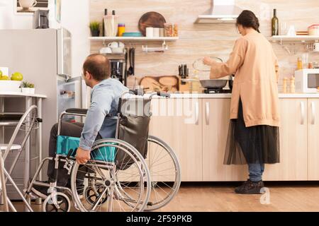 Behinderter Mann im Rollstuhl öffnet den Kühlschrank, während seine Frau kocht. Behinderter gelähmter behinderter Mann mit Gehbehinderung, der sich nach einem Unfall integriert. Stockfoto