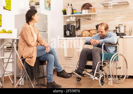 Frau schaut auf behinderten Mann im Rollstuhl, während das Essen auf Gasherd fertig ist. Behinderter gelähmter behinderter Mann mit Gehbehinderung, der nach einem Unfall integriert wird. Stockfoto