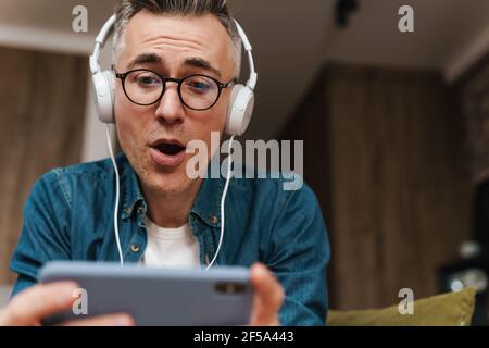 Überrascht Mann in Kopfhörer mit Handy, während sitzend auf Sofa zu Hause Stockfoto