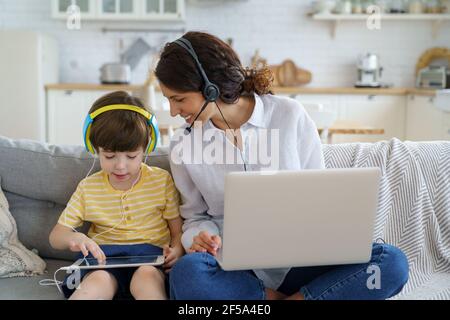 Glückliche Mutter mit Kind sitzt auf dem Sofa zu Hause während Lockdown Arbeit auf Laptop Kind spielen im Tablet Stockfoto