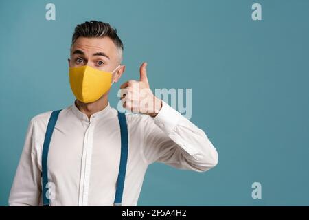 Aufgeregt grauhaarigen Mann in Gesichtsmaske zeigt Daumen nach oben isoliert Auf blauem Hintergrund Stockfoto