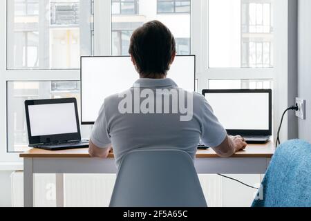 Ein Mann sitzt an einem Arbeitsplatz mit zwei Laptops und einem Monitor in der Nähe des Fensters. Remote-Arbeit von zu Hause in Quarantäne. Rückansicht Stockfoto