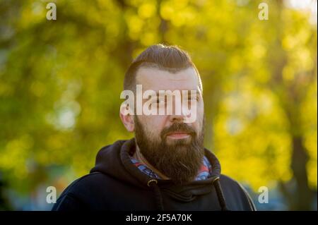 Ernst Kapuzen Kerl mit Bart sonnig im Freien auf verschwommenem Hintergrund. Mann mit Bart in schwarzem Hoodie Stockfoto