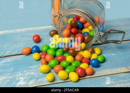 Bunte Süßigkeiten Kugeln im Glas gefallen aus auf blauem Holzhintergrund.Nahaufnahme der bunten Schokolade Süßigkeiten Hintergrund. Stockfoto