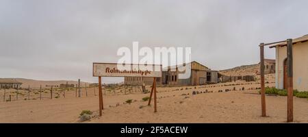 Das Schild Kolmannskuppe bei der Geisterstadt Kolmannskuppe In Namibia mit dem verlassenen Gebäude Panorama Stockfoto