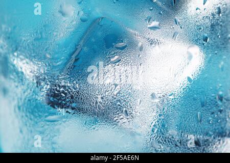 Eiswürfel in einem vernebelten Glas mit Tropfen Eiswasser Nahaufnahme Makro. Erfrischendes und kühlendes Getränk bei heißem Wetter. Blauer Hintergrund. Vollbild Stockfoto