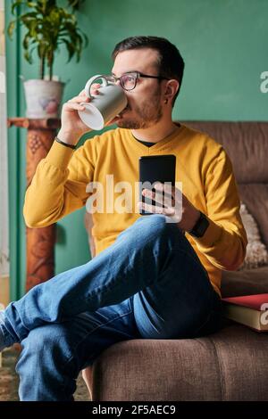 Ein junger Mann trinkt in seinem Leben eine Kaffeetasse Und nutzt sein Telefon Stockfoto